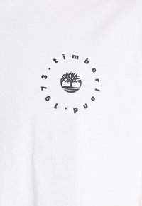 Camiseta de algodón blanca con un logo negro redondo que presenta un árbol y el texto "timberland 1973" a su alrededor. Textura suave, diseño casual.
