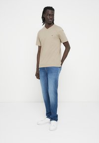 Ανοιχτό καφέ t-shirt με ντεκολτέ σε σχήμα V και κοντά μανίκια, σε συνδυασμό με ξεθωριαμένα μπλε τζιν και λευκά αθλητικά παπούτσια. Το μοντέλο στέκεται μπροστά σε απλό φόντο.