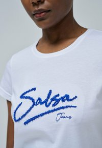 Camiseta de algodón blanca con bordado texturizado azul que dice "Salsa Jeans". Cuello redondo clásico y mangas cortas.