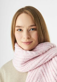 Jonge vrouw met lichtbruin haar en blauwe ogen, gekleed in een zachte, dikke roze gebreide sjaal en een beige top tegen een witte achtergrond.