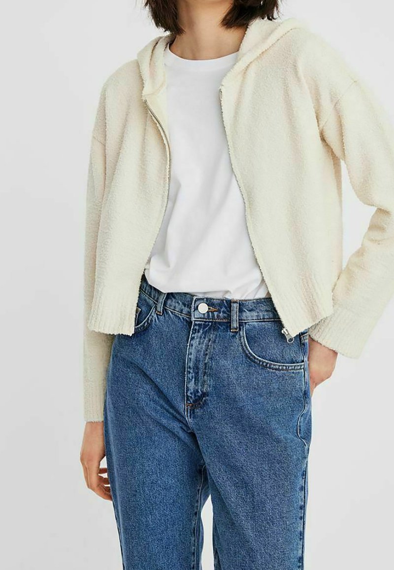 Sweat à capuche court de couleur crème en tissu doux, avec une fermeture éclair et une capuche attachée, associé à un jean bleu taille haute.