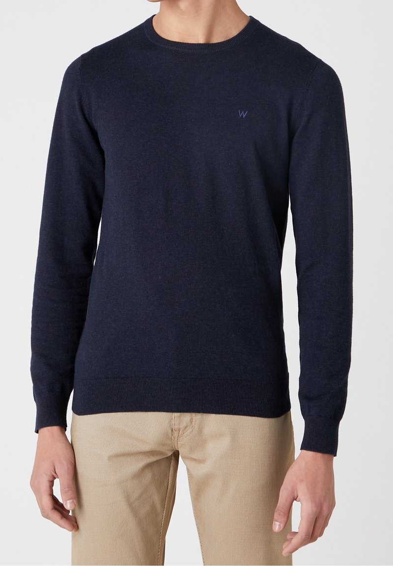 Homme portant un pull bleu marine col rond à manches longues et un pantalon beige sur un fond uni.