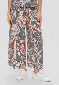 Femme portant un pantalon large à motifs floraux abstraits multicolores et des baskets blanches, debout devant un fond clair.