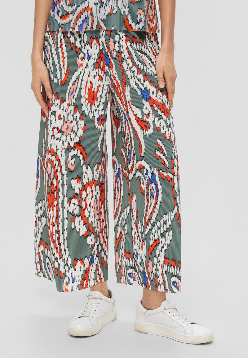 Femme portant un pantalon large à motifs floraux abstraits multicolores et des baskets blanches, debout devant un fond clair.