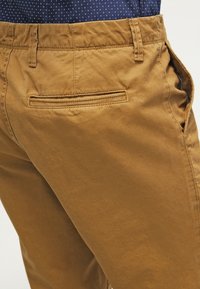 Pantalon en coton marron avec poche passepoilée à l'arrière, passants pour ceinture et poche latérale portée avec une chemise bleu marine à motifs.