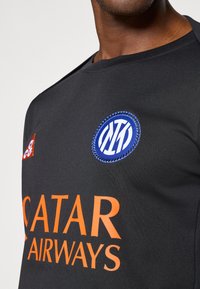 Μαύρη αθλητική φανέλα με μπλε κυκλικό λογότυπο με λευκά γράμματα, πορτοκαλί κείμενο "QATAR AIRWAYS" και ένα μικρό κόκκινο λογότυπο στον αριστερό ώμο.