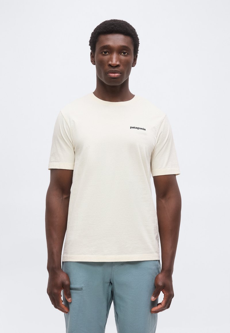 Hombre vistiendo una camiseta Patagonia de color blanco roto lisa y pantalones azul claro, de pie frente a un fondo gris claro, mirando al frente.