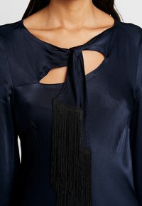 Blusa de satén navy con escote recortado, detalle de lazo y decoración de flecos negros en cascada en el frente. Textura suave.