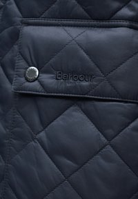 Μπλε σκούρο καπιτονέ μπουφάν με εμφανές κουμπί πιέτας και κεντημένο λογότυπο "Barbour" στην μπροστινή τσέπη. Ομαλή υφή και γεωμετρικό σχέδιο.