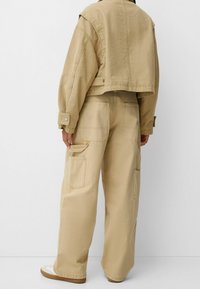 Chaqueta beige corta con un ajuste oversize, mangas largas y puños abotonados, combinada con pantalones beige de pierna ancha que cuentan con múltiples bolsillos tipo cargo.