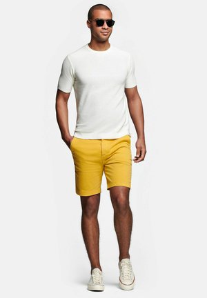 Man met zwarte zonnebril, wit T-shirt met korte mouwen, gele korte broek en witte sneakers, lopend met één hand in zijn zak.