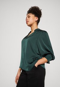 Blouse en satin vert foncé avec un décolleté en V et des manches bouffantes, présentant une texture lisse et une coupe décontractée, associée à un pantalon noir.