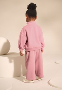 Rožinis vilnos džemperis su aukšta apykakle ir riboto audinio rankogaliais, derinamas su plataus silueto laisvalaikio kelnėmis. Modelis avėdamas baltus sportinius batelius.