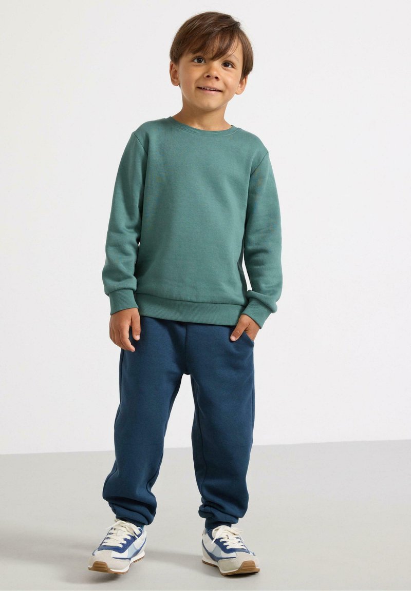 Felpa verde con polsini a coste, abbinata a pantaloni della tuta blu con tasche laterali. Sneakers bianche e blu con suole beige.