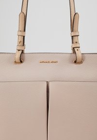 Beżowa skórzana torba tote z dwoma przegrodami, złotymi detalami i podwójnymi regulowanymi paskami; posiada wytłoczone logo "Michael Kors".