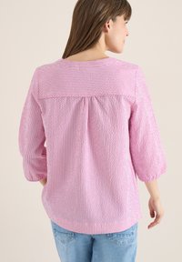 Mujer con blusa de manga larga a rayas rosas y blancas con detalle fruncido en la espalda, combinada con vaqueros azul claro, de espaldas.