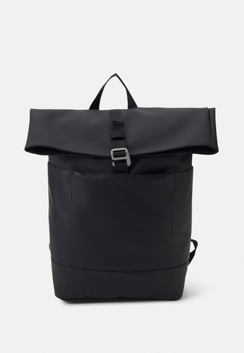 Sac à dos noir à fermeture roulante avec poche avant et boucle argentée sur un fond clair uni.