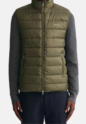 Uomo che indossa un gilet trapuntato verde oliva con cerniera sopra un maglione grigio a maniche lunghe e pantaloni scuri, in piedi con le braccia rilassate ai lati.