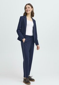 Námornícky modrý blazer a zhodné jogger nohavice majú voľný strih, biele bočné pruhy a elastický pás s šnúrkou. Skombinované s bielym topom a tmavými topánkami.