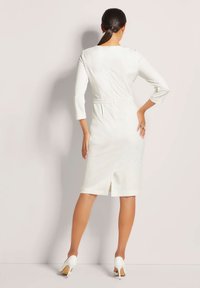 White fitted dress with three-quarter sleeves, back slit, and textured fabric, paired with white high-heeled shoes, seen from the back.