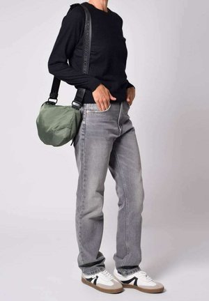 Groene gewatteerde tas met verstelbare schouderband, zwarte lange mouwen trui, grijze recht afgewerkte jeans en witte sneakers met bruine accenten.