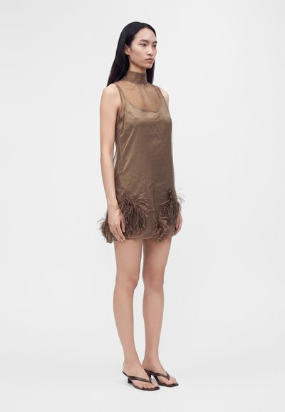 Femme aux longs cheveux noirs portant une robe marron sans manches avec des détails en plumes près de l'ourlet et des sandales marron à talons bas, debout sur fond blanc.
