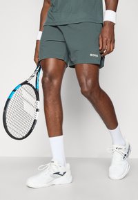 Βραχέα αθλητικά σορτς σε σκούρο γκρι, με λογότυπο στο τελείωμα, συνδυασμένα με λευκά αθλητικά παπούτσια και ρακέτα τένις.