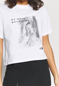 Camiseta blanca recortada con una ilustración gráfica negra de manos en posición de oración y el texto "MOVEMOS MONTAÑAS." Material de algodón suave.