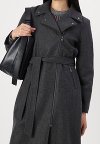 Grå uldblandingsfrakke med lynlås foran, sidelommer og bælte. Har en klippeskrå krave og trykknapdetaljer. Følgende med en sort taske.