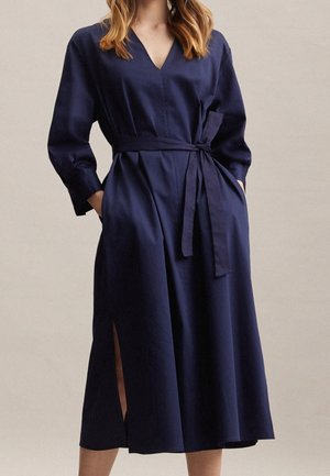 Kobieta ubrana w granatową, długorękawową sukienkę do połowy łydki z dekoltem w serek, rozcięciem z boku i paskiem wiązanym w talii, z rękami w kieszeniach.