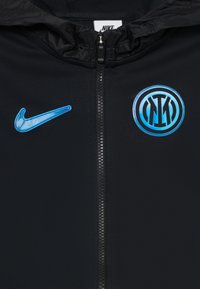 Čierna bunda z prednej strany s modrým logom Nike swoosh na ľavom prsníku a emblémom futbalového klubu Inter Miláno na pravom prsníku, so zipsom uprostred.