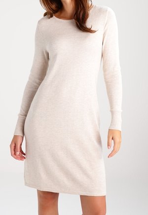 Kobieta ubrana w jasnobeżową, długorękawową dzianinową sukienkę sięgającą powyżej kolan na jednolitym tle.