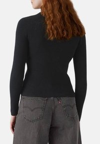 Top lungo a maniche nere a costine con silhouette aderente, indossato con jeans grigi a vita alta con tasche posteriori e una etichetta rossa.