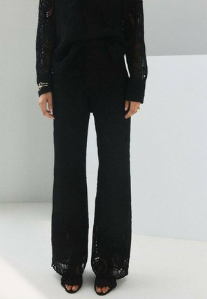 Frau trägt ein langärmeliges Oberteil aus schwarzer Spitze und passende weit geschnittene Spitzenhosen, steht vor einem weißen und grauen Hintergrund.