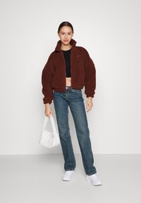 Brun, kort, zip-up fleecejakke med høy krage, kombinert med høyhylte blå jeans og hvite sneakers. Holder en liten hvit veske.