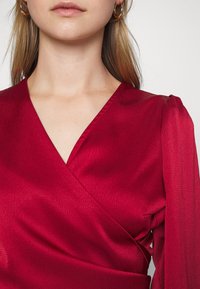 Robe portefeuille rouge avec une finition texturée, décolleté en V et manches longues semi-transparentes. Présente des plis subtils et une silhouette ajustée.