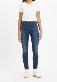 Jeans de denim oscuros de tiro alto y corte ajustado. Combinados con una camiseta blanca con un pequeño logo rojo. Zapatillas blancas con suelas beige.