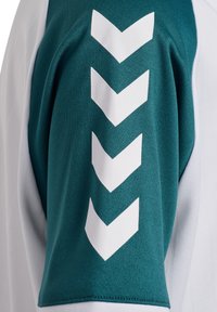 Kortærmet sportstrøje med tealfarvet stof med en struktureret finish, hvid chevron-design på ærmet og en kontrasterende hvid krop.