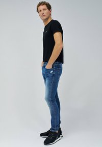 Camiseta negra ajustada, jeans azules desgastados y zapatillas deportivas negras. El conjunto presenta un diseño sencillo y un aspecto casual.