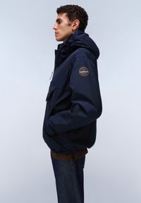 Junger Mann im Profil, der eine marineblaue Kapuzenjacke mit rundem Aufnäher am Ärmel und dunkelblaue Jeans vor einem einfarbigen Hintergrund trägt.