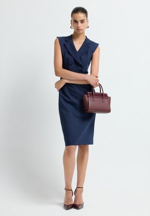 Donna che indossa un vestito a tubino senza maniche di colore blu navy, tiene in mano una borsa bordeaux e indossa scarpe col tacco a punta bordeaux abbinate e orecchini a cerchio dorati.
