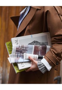 Trench-coat marron avec manches retroussées, tenant des magazines verts, blancs et gris, à côté d'une petite bouteille blanche avec un bouchon noir.