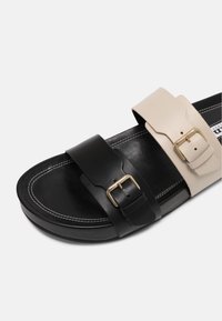 Mule noire avec deux brides réglables, une beige et une noire, chacune dotée de boucles dorées et d'une couture blanche autour de la semelle.