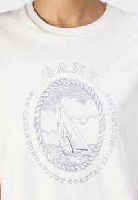 Hvid bomulds t-shirt med et blåt trykt design af en sejlbåd inden for en reboval. Teksten inkluderer "GANT" og "NEW HAVEN CONNECTICUT COASTAL SAILING CLUB 1949."