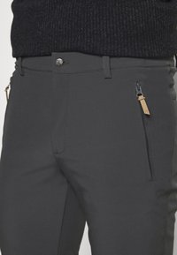 Un pantalon gris au toucher lisse dispose d'une poche zippée sur le côté, d'un matériel en métal à la taille et de bandes latérales contrastantes.