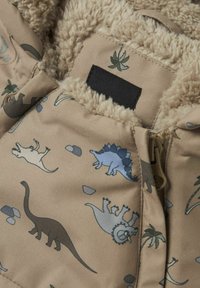Tissu beige avec des motifs de dinosaures en bleu pâle, vert et gris. Doublure intérieure douce et duveteuse et une étiquette noire près de la fermeture éclair.