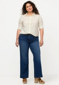 Camisa de botón beige claro con mangas remangadas, combinada con jeans de pierna ancha azul oscuro y sandalias de cuña marrones. Tejido suave con un corte casual.