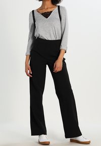 Graues Langarmoberteil mit V-Ausschnitt und Spitzenakzent, kombiniert mit schwarzen weit geschnittenen Hosen und weißen Sneakers mit braunen Sohlen.