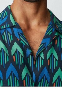 Lyhythihainen paita, jossa tummansininen tausta ja vihreän, sinisen sekä oranssin geometrinen kuvio. Kangas vaikuttaa kevyeltä ja sileäpintaiselta.