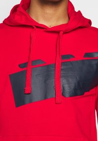 Czerwona bluza z kapturem z dużym wytłoczonym czarnym logo na piersi, z sznurkami i teksturowaną tkaniną. Kieszeń typu kangur z przodu.